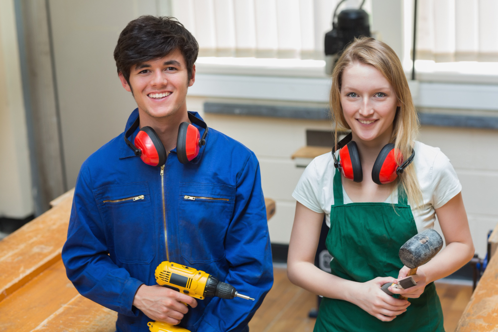 Two smiling vocational students, one male with a drill and one female with a hammer, in a workshop for plumber courses near me